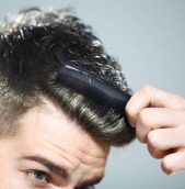 Mid 20's hipster man combing his hair.Using small black comb.He has neat hairstyle with very short hair on the side and one inch length on top,dark brown.Image cut just below his eyes.Gray background.